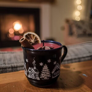 Handgemachte Glühwein Duftkerze in dekorativem Winterbecher mit Weihnachtsmotiv, garniert mit Zimtstange, Sternanis und Orangenscheibe vor gemütlichem Kamin mit Lichterketten